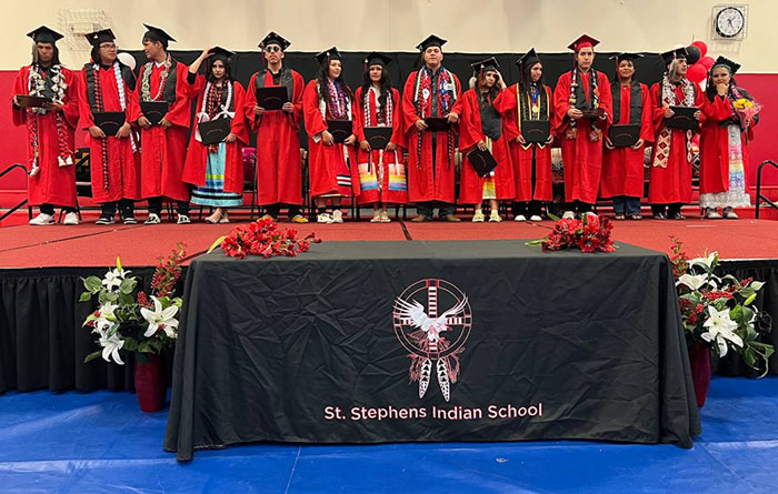 Graduates in red caps and gowns stand on a stage during a ceremony at St Stephens, with floral decorations and a school banner in front.