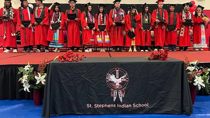 Graduates in red caps and gowns stand on a stage during a ceremony at St Stephens, with floral decorations and a school banner in front.